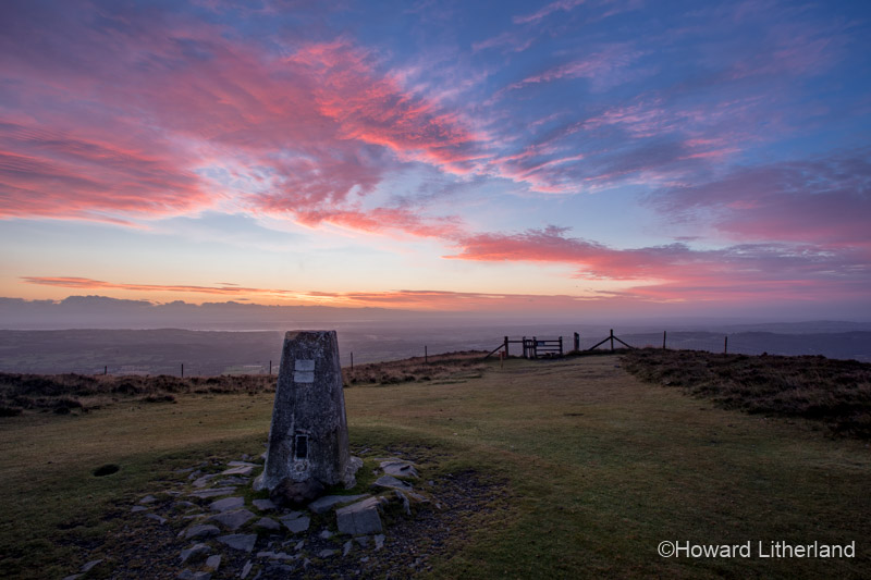 Clouds lit by dawn light over an Ordinance Survey trig point at the summit of Moel Famau, North Wales
