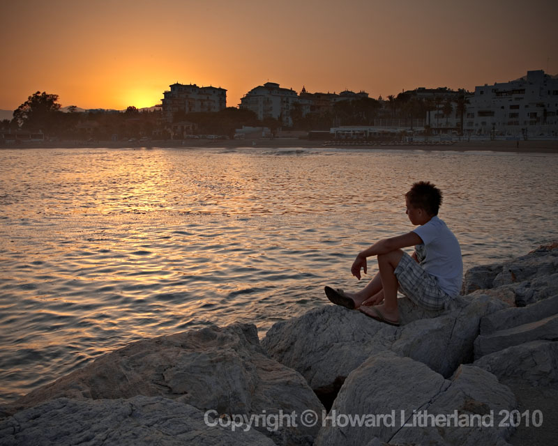 Boy sitting on harbour wall at sunset, Marbella, Spain