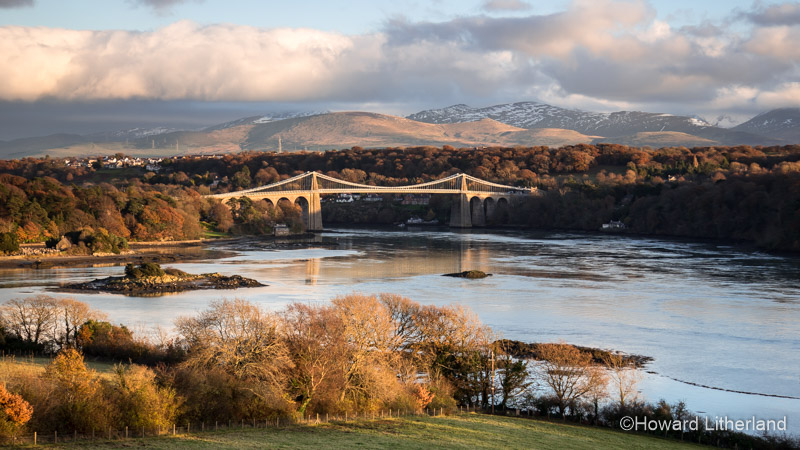 Thomas Telford's iconic suspension bridge over the Menai Straits, North Wales, lit by a low afternoon sun with snow capped mountains in the background