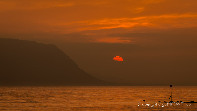 The setting sun, partially obscured by a line of cloud next to the headland at Penmaenmawr, viewed from Deganwy on the North Wales coast